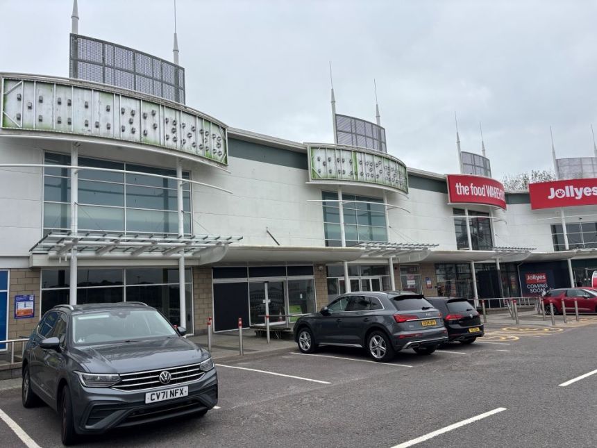 The exterior of Parc Fforestfach retail park showing the vacant units 4 and 5 in the foreground, with The Food Warehouse and a Jollyes "coming soon" sign visible to the right, and cars parked in the foreground.