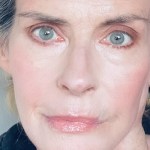 A close-up selfie of Annabel Schofield in later life, showing her striking blue-grey eyes and blonde-grey hair pulled back, wearing a black top.