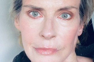 A close-up selfie of Annabel Schofield in later life, showing her striking blue-grey eyes and blonde-grey hair pulled back, wearing a black top.