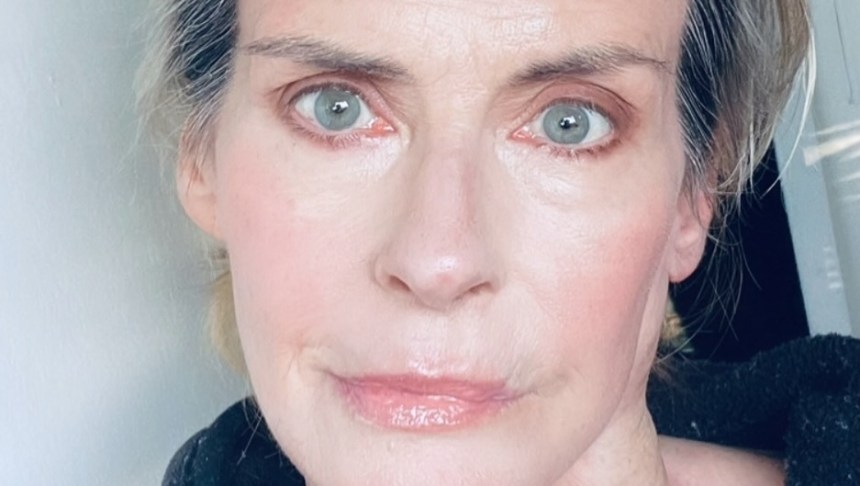 A close-up selfie of Annabel Schofield in later life, showing her striking blue-grey eyes and blonde-grey hair pulled back, wearing a black top.