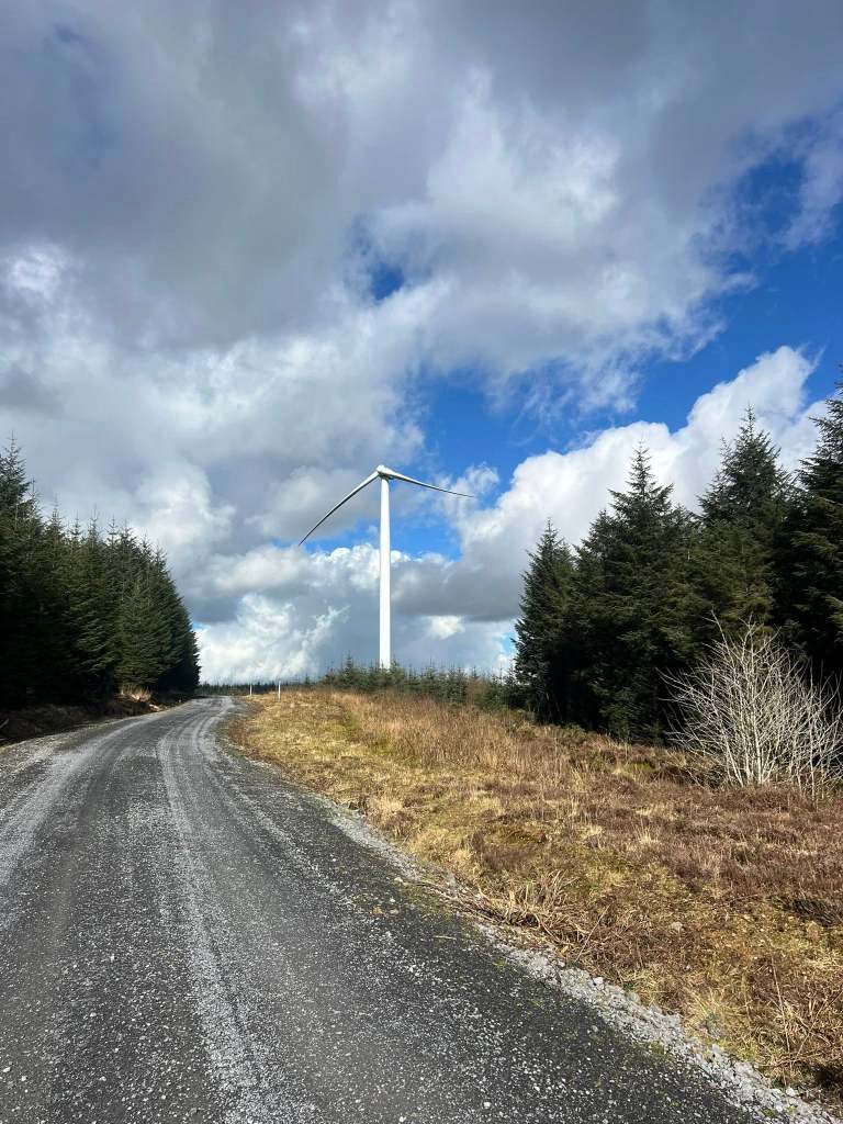 A tall white wind turbine standing in a forest clearing against a cloudy sky, with only two of its three blades attached
