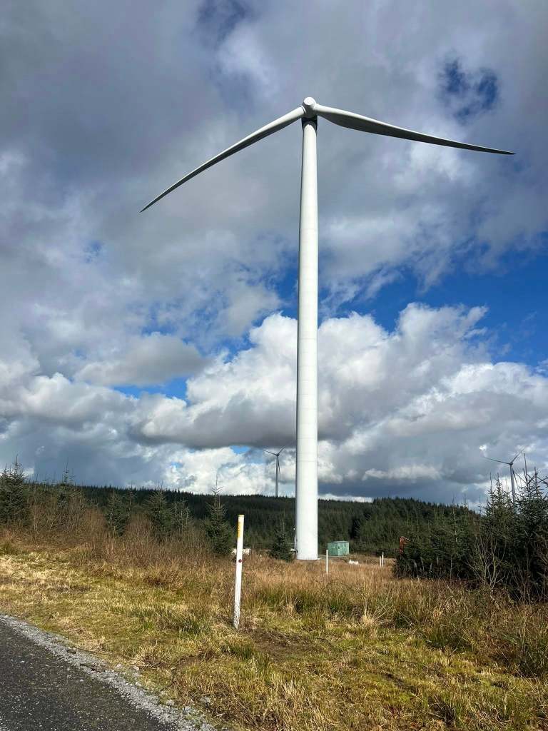 A tall white wind turbine standing in a forest clearing against a cloudy sky, with only two of its three blades attached
