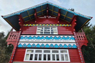 A photo of the ornate, red-painted Swiss Cottage in Singleton Park, showing its decorative balconies and inscriptions under a cloudy sky.