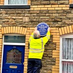 A workman in a high-visibility yellow jacket is seen from behind as he carefully fixes the blue Ray Milland plaque onto the wall of a terraced house.