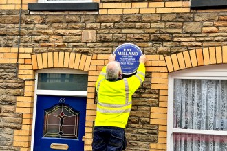 A workman in a high-visibility yellow jacket is seen from behind as he carefully fixes the blue Ray Milland plaque onto the wall of a terraced house.