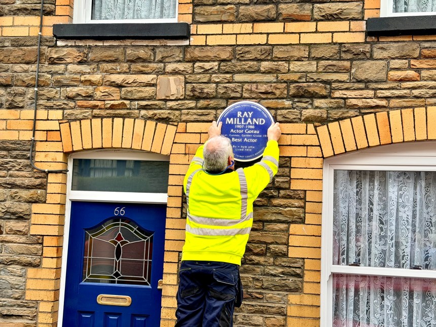 A workman in a high-visibility yellow jacket is seen from behind as he carefully fixes the blue Ray Milland plaque onto the wall of a terraced house.