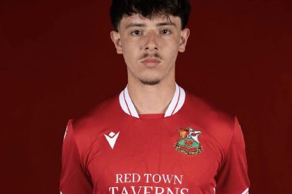 A young male footballer, James Lester, wearing a red Llanelli Town AFC kit with "RED TOWN TAVERNS" sponsored on the front, stands against a plain red background, looking directly at the camera.