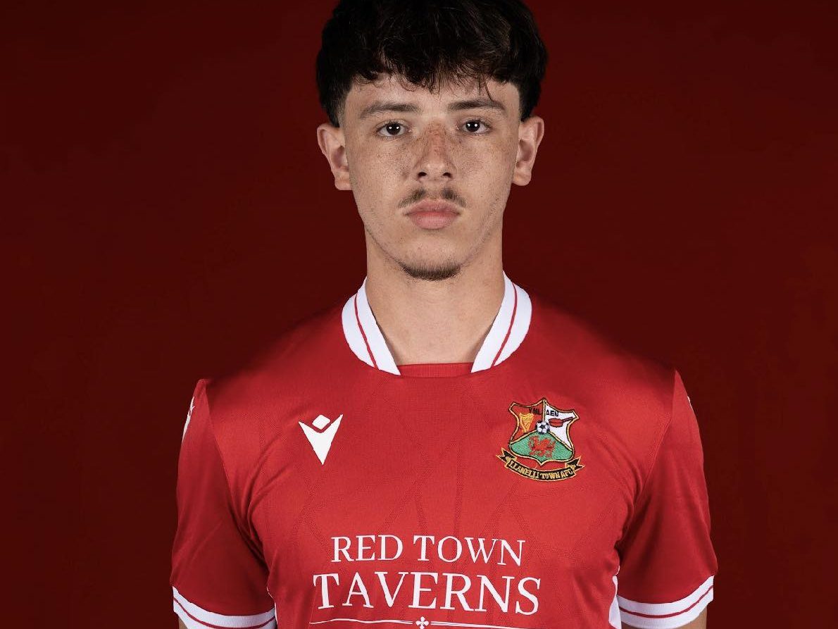 A young male footballer, James Lester, wearing a red Llanelli Town AFC kit with "RED TOWN TAVERNS" sponsored on the front, stands against a plain red background, looking directly at the camera.