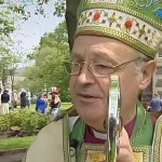Anthony Pierce, dressed in full bishop's vestments including a green and gold mitre and holding a ceremonial staff, stands outside Swansea Minster. Two individuals in the background have been blurred.