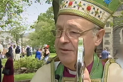 Anthony Pierce, dressed in full bishop's vestments including a green and gold mitre and holding a ceremonial staff, stands outside Swansea Minster. Two individuals in the background have been blurred.