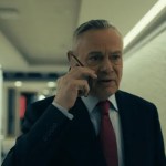 Martin Clunes in character as former BBC newsreader Huw Edwards in a television drama, wearing a suit and tie against a studio-style background.