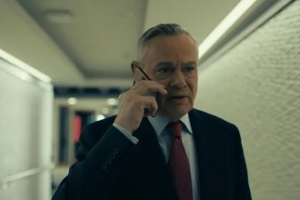 Martin Clunes in character as former BBC newsreader Huw Edwards in a television drama, wearing a suit and tie against a studio-style background.