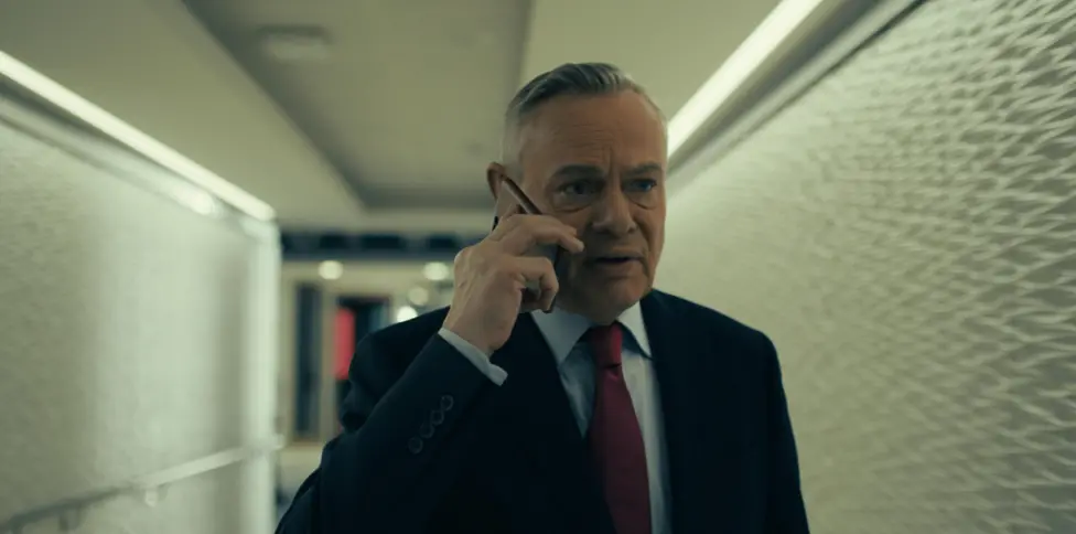 Martin Clunes in character as former BBC newsreader Huw Edwards in a television drama, wearing a suit and tie against a studio-style background.