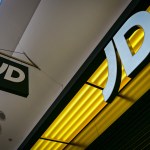 A close-up, low-angle shot of the glowing white "JD" logo on a bright yellow background, with a smaller black and white JD sign hanging nearby.