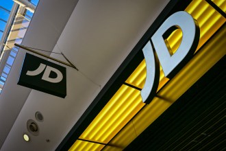 A close-up, low-angle shot of the glowing white "JD" logo on a bright yellow background, with a smaller black and white JD sign hanging nearby.