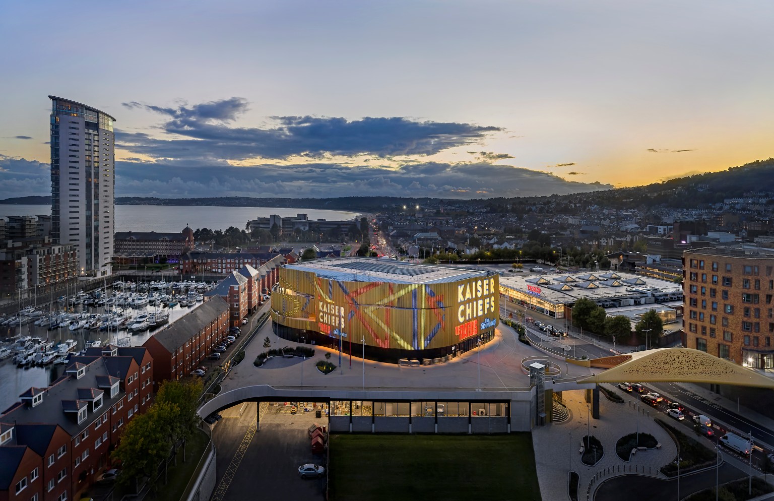 An aerial view of the modern Swansea Building Society Arena building at dusk, with its illuminated facade, surrounded by other city buildings and the bay in the background.