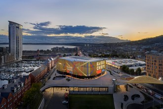 An aerial view of the modern Swansea Building Society Arena building at dusk, with its illuminated facade, surrounded by other city buildings and the bay in the background.