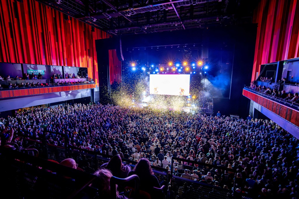 A wide shot of a large, enthusiastic crowd inside a concert arena, with a stage illuminated by bright lights and confetti falling from the ceiling.
