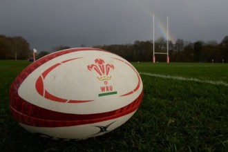 A close-up of a white and red rugby ball with the WRU three-feathers logo on a green grass pitch, with rugby posts and a rainbow in the background.