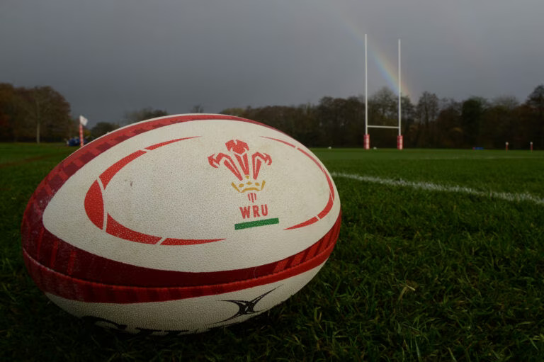 A close-up of a white and red rugby ball with the WRU three-feathers logo on a green grass pitch, with rugby posts and a rainbow in the background.
