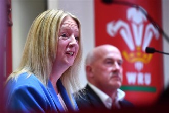 Abi Tierney speaking at a press conference in a blue jacket, with a blurred man in a suit and the WRU logo in the background.