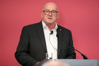 A portrait of Richard Collier-Keywood, a man with a bald head and glasses, wearing a dark suit and white shirt, speaking at a podium against a red background.