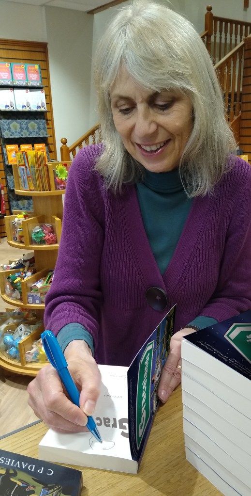 Author C.P. Davies signing copies of her book Gracelands at a bookshop, seated at a table with stacks of books.
