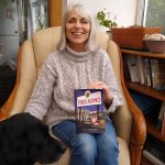 Author C.P. Davies sitting in an armchair holding her book Trecadno with a black dog beside her in a cosy room.