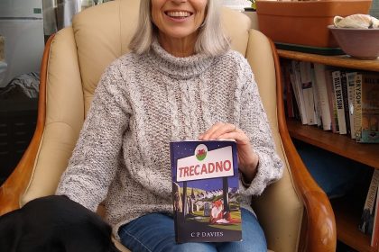 Author C.P. Davies sitting in an armchair holding her book Trecadno with a black dog beside her in a cosy room.
