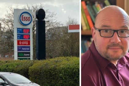 Esso fuel station sign showing unleaded at 153.9p and diesel at 180.9p, with a 30mph sign nearby. Beside it, a portrait of Carl Peters‑Bond indoors in front of bookshelves.