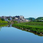 A view of Carmarthen town from Lesneven Bridge, showing the River Tywi in the foreground with green marshland on the right bank, and the town's buildings and surrounding hills under a clear blue sky.
