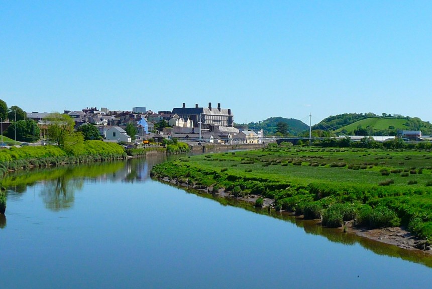 A view of Carmarthen town from Lesneven Bridge, showing the River Tywi in the foreground with green marshland on the right bank, and the town's buildings and surrounding hills under a clear blue sky.