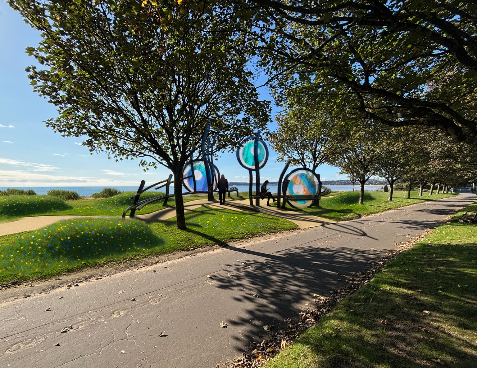 An artist's impression of a public memorial on a grassy area next to a paved path and trees. The memorial consists of three large, circular glass panels with blue and orange peacock-butterfly-inspired designs, held up by dark, curved metal frames that also form seating. In the background, people are walking on the path, and the sea is visible under a blue sky.