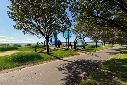 An artist's impression of a public memorial on a grassy area next to a paved path and trees. The memorial consists of three large, circular glass panels with blue and orange peacock-butterfly-inspired designs, held up by dark, curved metal frames that also form seating. In the background, people are walking on the path, and the sea is visible under a blue sky.