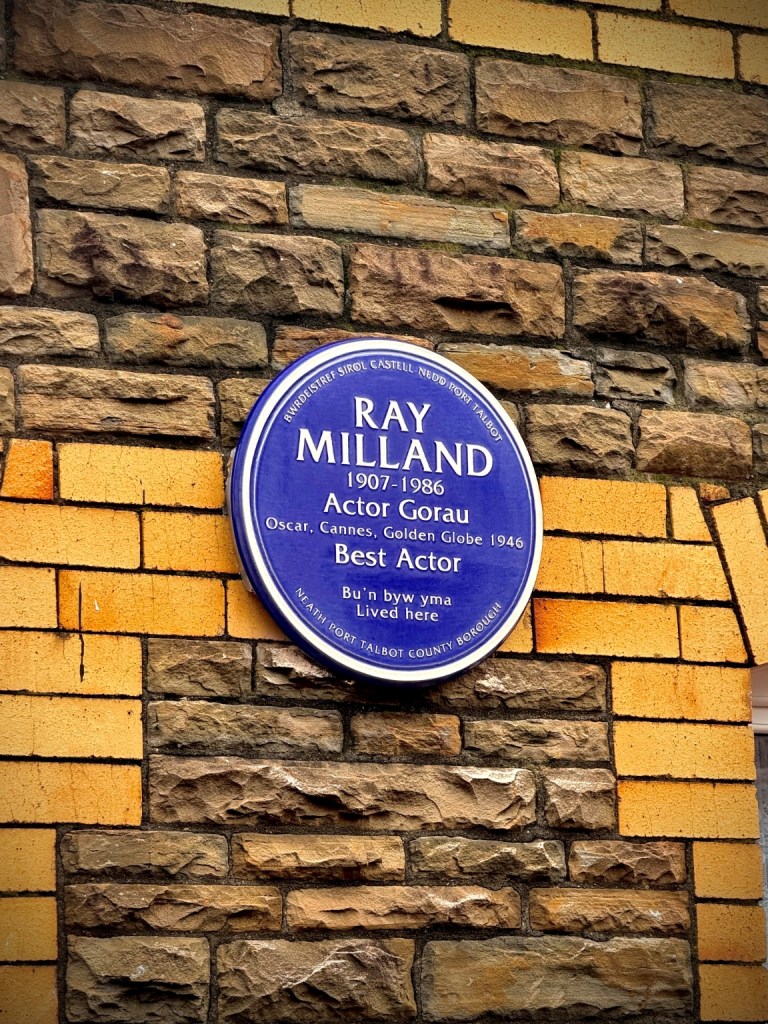 A close-up shot of the blue circular plaque for Ray Milland installed on a stone and brick wall. It lists his name, dates, and major awards.