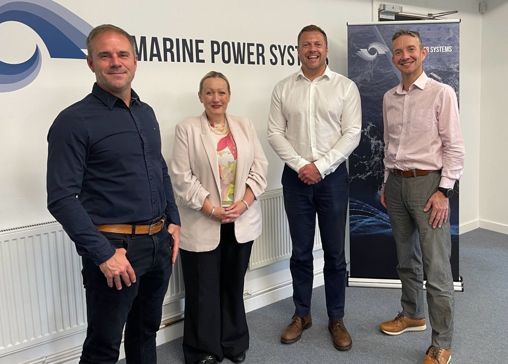 Four people standing in an office in front of a wall with the Marine Power Systems logo. From left to right: a man in a black shirt, a woman in a cream jacket, a man in a white shirt, and a man in a pink shirt.
