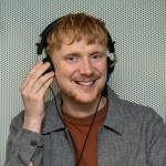 A man wearing headphones smiles while adjusting them during a hearing test inside a soundproof booth.