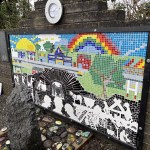 The finished mosaic memorial, a long rectangular artwork mounted on a brick wall outdoors. The top half is a colourful landscape with a bright sun, while the bottom half is a black and white scene depicting the mining disaster.