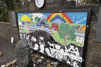 The finished mosaic memorial, a long rectangular artwork mounted on a brick wall outdoors. The top half is a colourful landscape with a bright sun, while the bottom half is a black and white scene depicting the mining disaster.