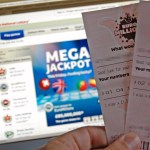 A close-up photo of a hand holding two EuroMillions lottery tickets in front of a computer screen displaying the National Lottery website with a "Mega Jackpot" banner.