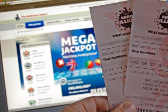 A close-up photo of a hand holding two EuroMillions lottery tickets in front of a computer screen displaying the National Lottery website with a "Mega Jackpot" banner.