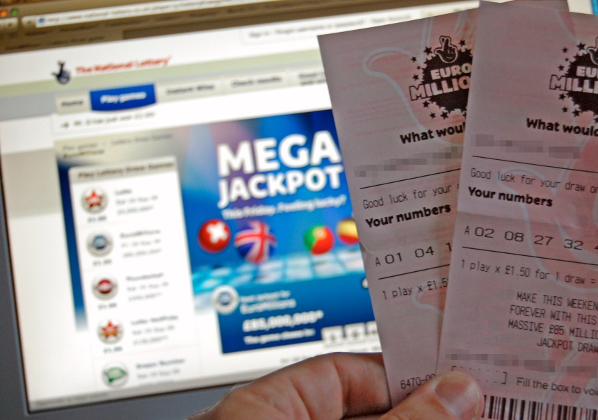 A close-up photo of a hand holding two EuroMillions lottery tickets in front of a computer screen displaying the National Lottery website with a "Mega Jackpot" banner.