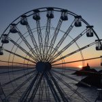 A large white big wheel is silhouetted against a sunset over the sea, with the dark outline of a headland and lighthouse visible in the distance.