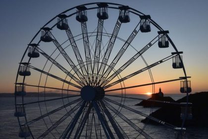 A large white big wheel is silhouetted against a sunset over the sea, with the dark outline of a headland and lighthouse visible in the distance.