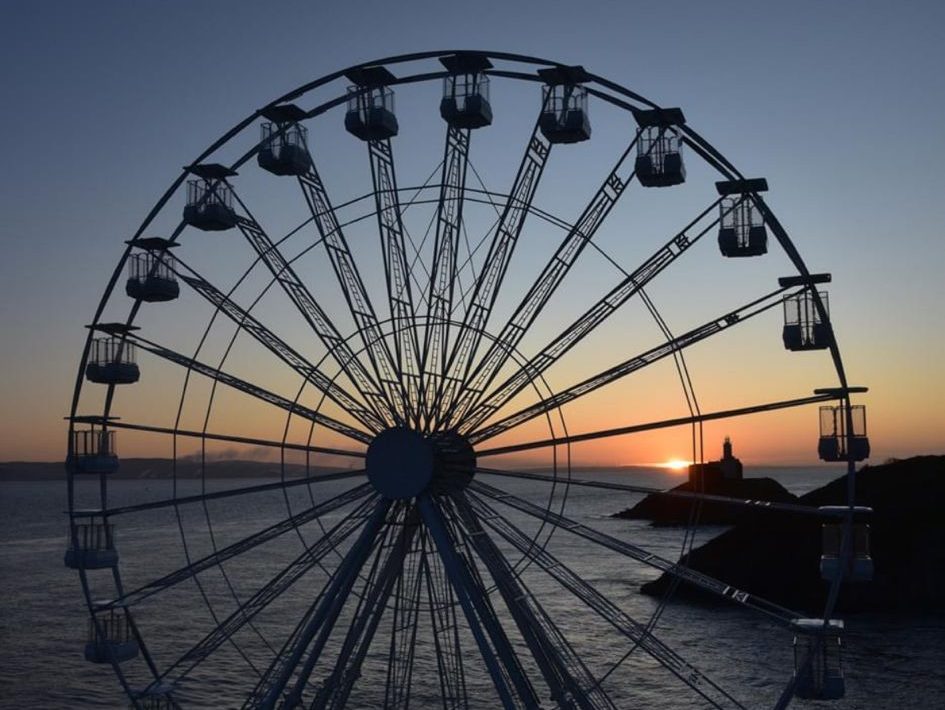 A large white big wheel is silhouetted against a sunset over the sea, with the dark outline of a headland and lighthouse visible in the distance.