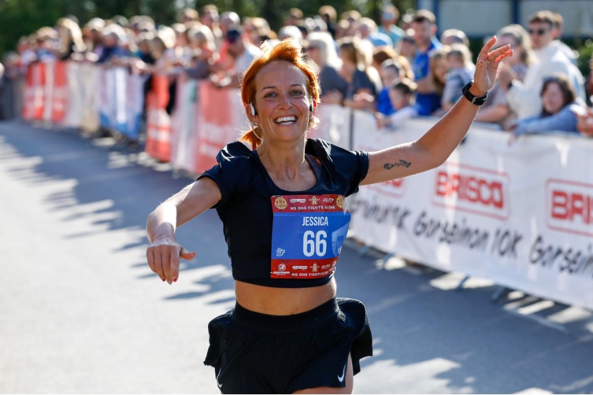 Female runner wearing race number 66 raises her arms while finishing the Gorseinon 10K, with spectators lining the barriers behind her.