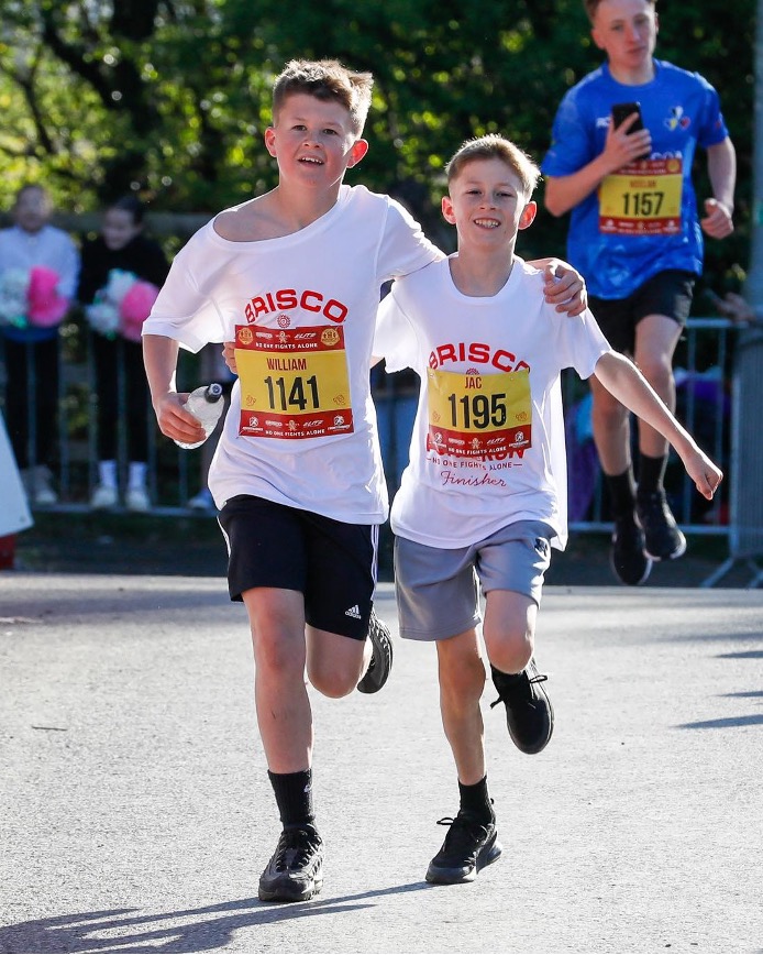 Two boys wearing race bibs run side by side and smile as they approach the finish line during a community fun run.