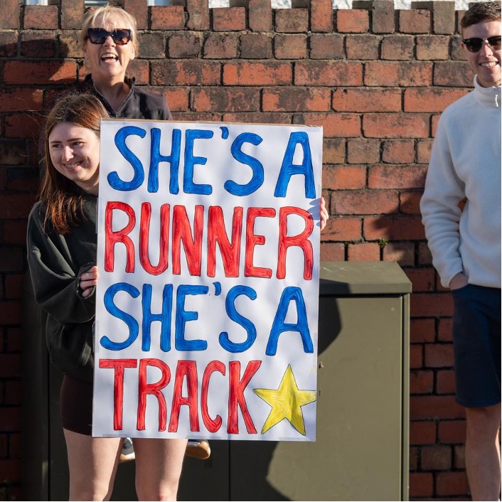 Spectator holding a handmade sign reading “She’s a runner she’s a track star” while watching runners pass during a road race.