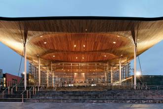 A wide-angle exterior shot of the Senedd building in Cardiff Bay at twilight, showing its massive glass walls and the iconic, illuminated undulating wooden ceiling supported by slender columns.
