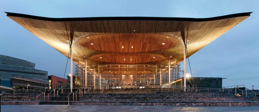 A wide-angle exterior shot of the Senedd building in Cardiff Bay at twilight, showing its massive glass walls and the iconic, illuminated undulating wooden ceiling supported by slender columns.